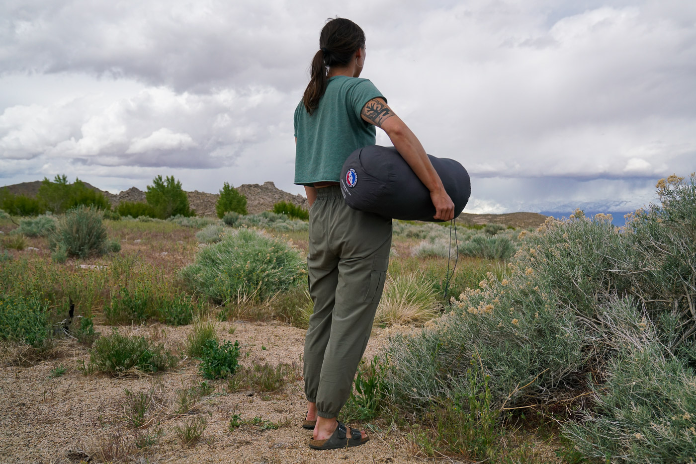A person with their back to the camera holds a sleeping bag in a stuff sack under their arm.