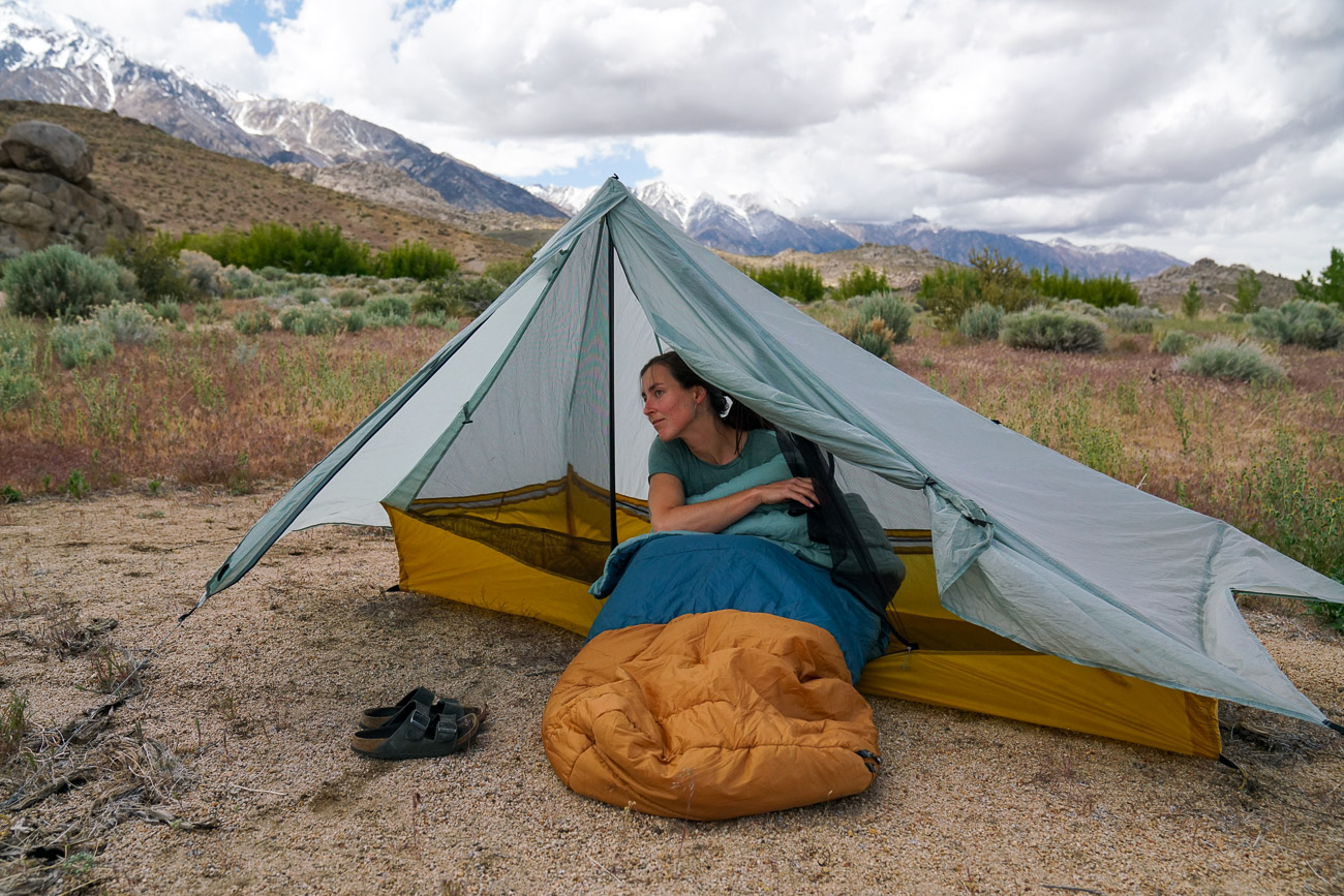 A person sitting in a sleeping bag looks out from inside a tent. There are mountains in the background.