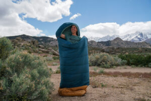 A person stands inside a sleeping bag in front of mountains.
