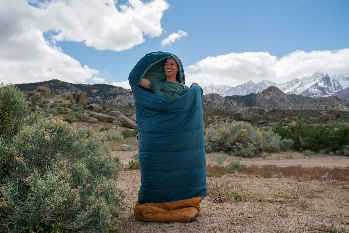 A person stands inside a sleeping bag in front of mountains.
