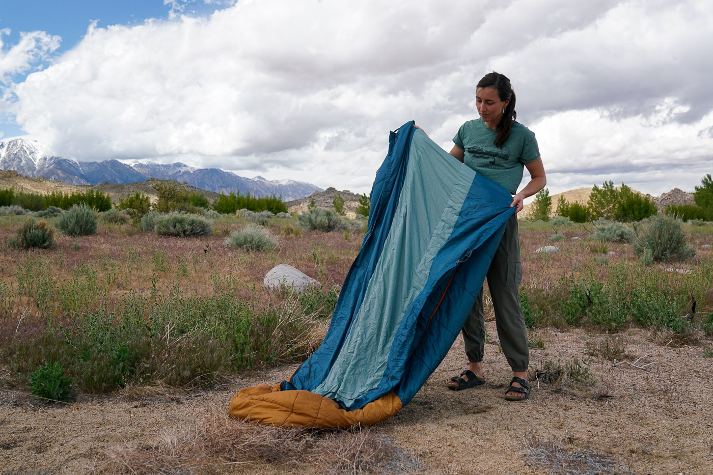 A person stands holding up a sleeping bag. There are mountains in the background.