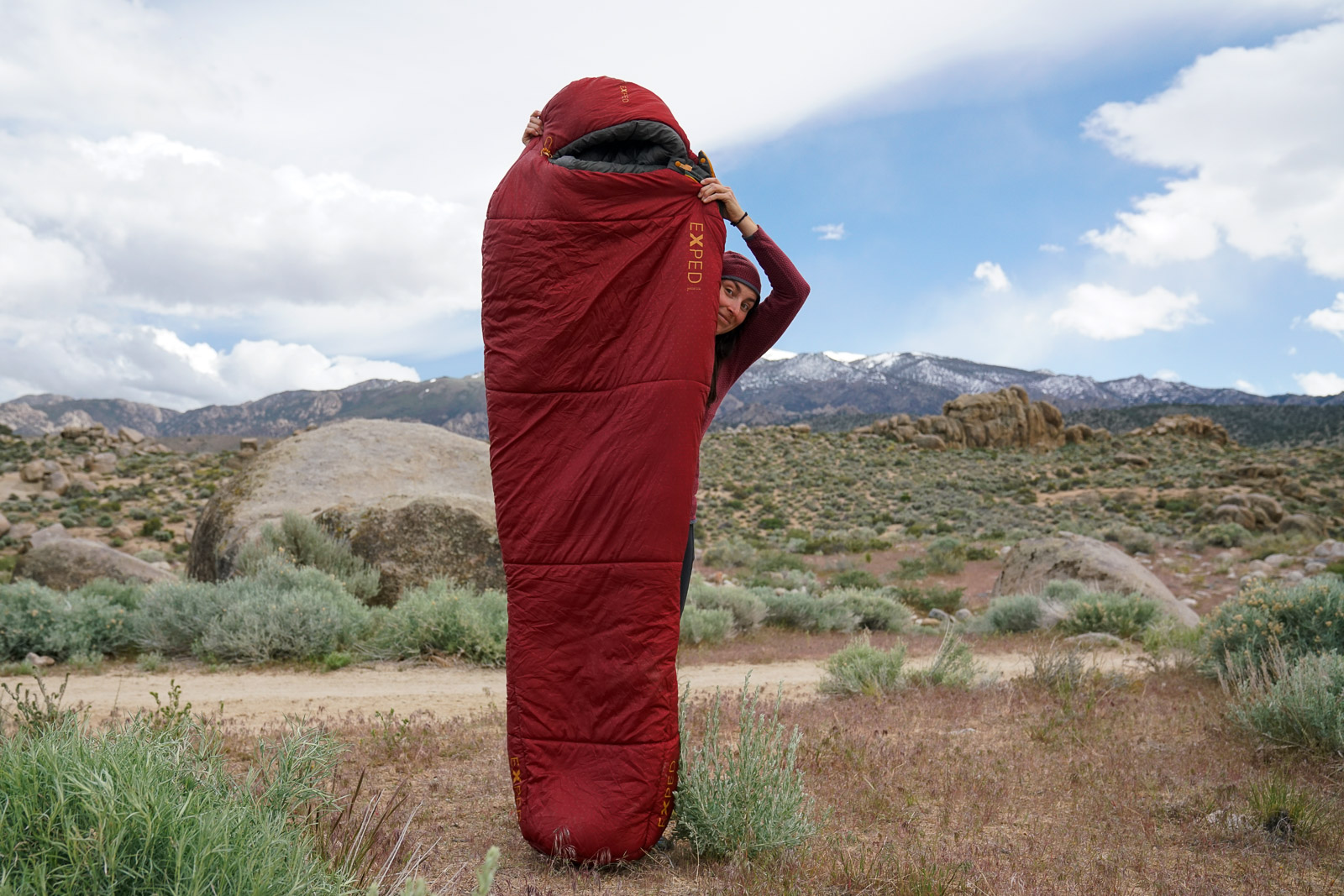 A person stands holding up a sleeping bag in front of their body. There are boulders in the background.