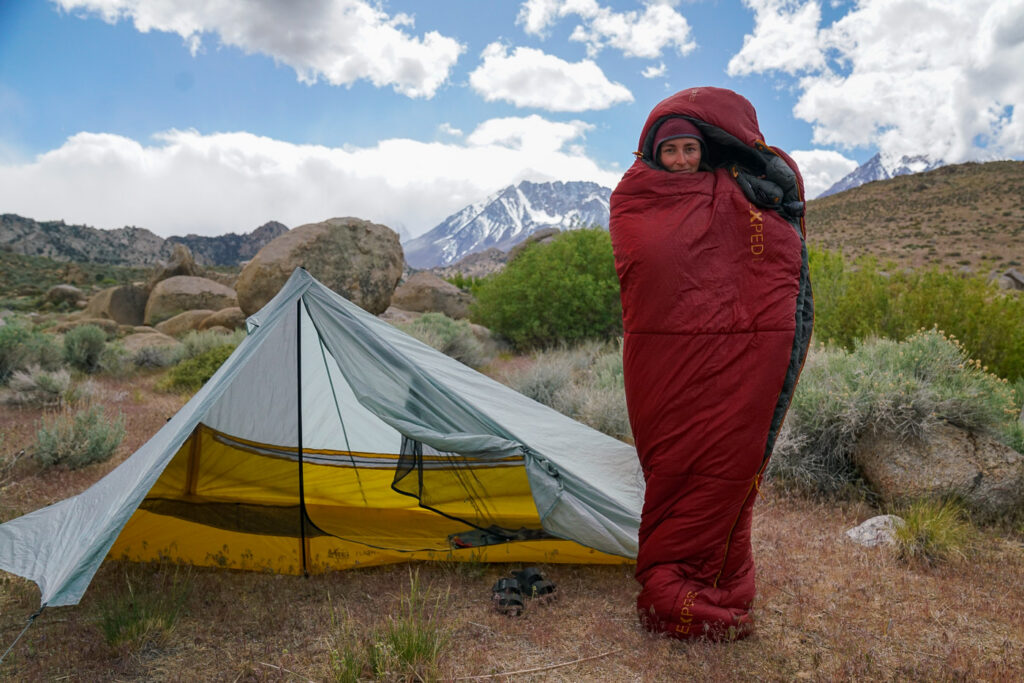 A person wrapped in a sleeping bag stands next to a tent with mountains in the background.