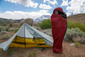 A person wrapped in a sleeping bag stands next to a tent with mountains in the background.
