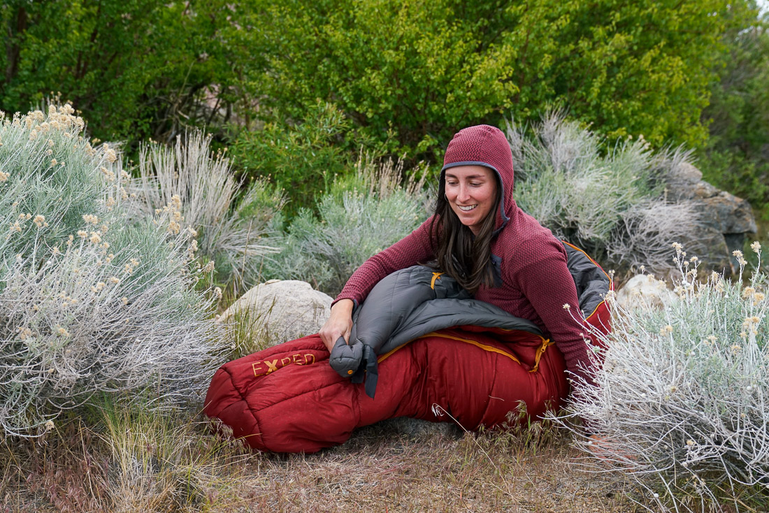 A person sits in a sleeping bag on the ground.