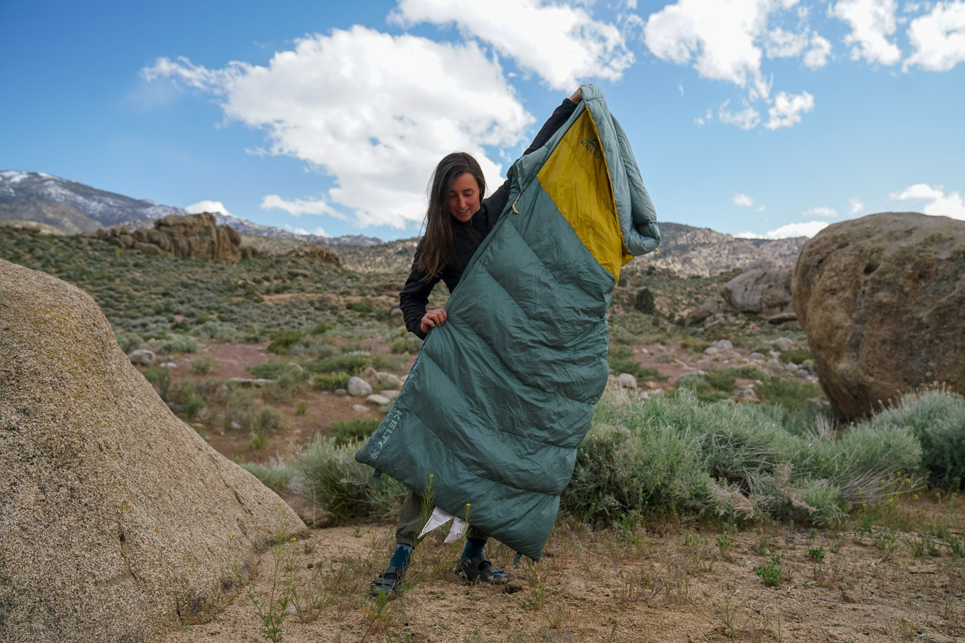 A person stands holding a sleeping bag across their body. There are boulders in the background.