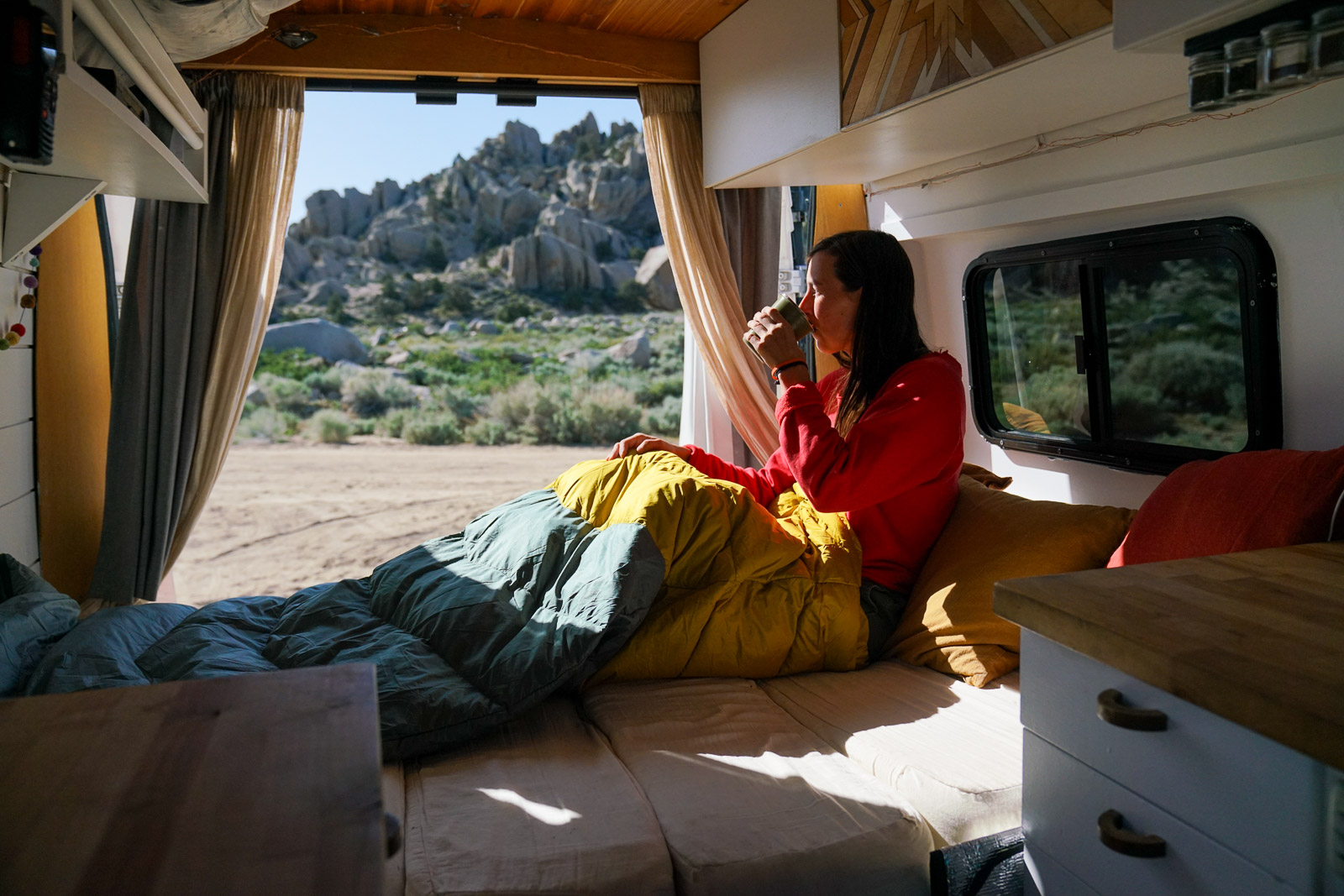 A person drinks coffee sitting in a sleeping bag on the bed of a camper van. The van's doors are open revealing boulders in the background.