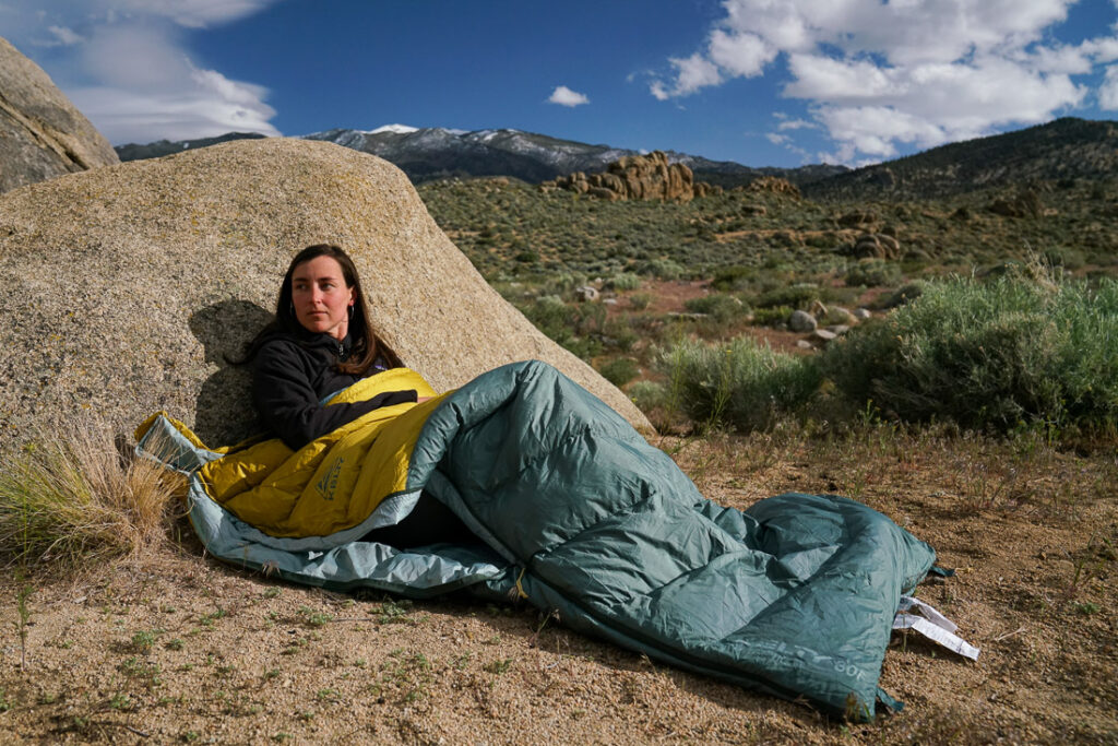 A person leans against a boulder while sitting in a sleeping bag. There are boulders in the background.