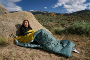 A person leans against a boulder while sitting in a sleeping bag. There are boulders in the background.