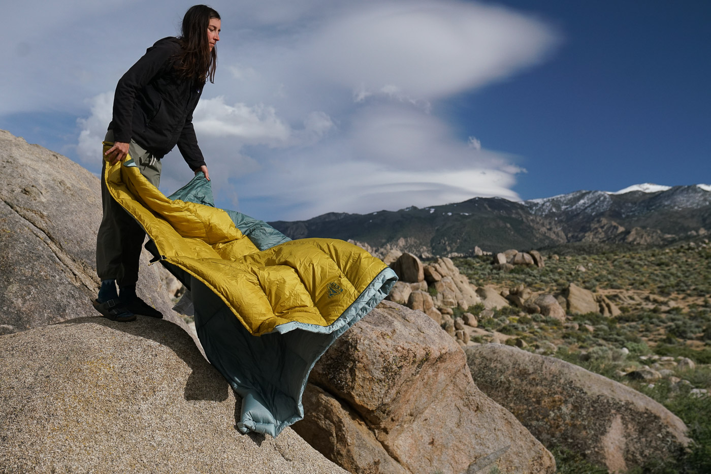 A person stands on a boulder and shakes out a sleeping bag.