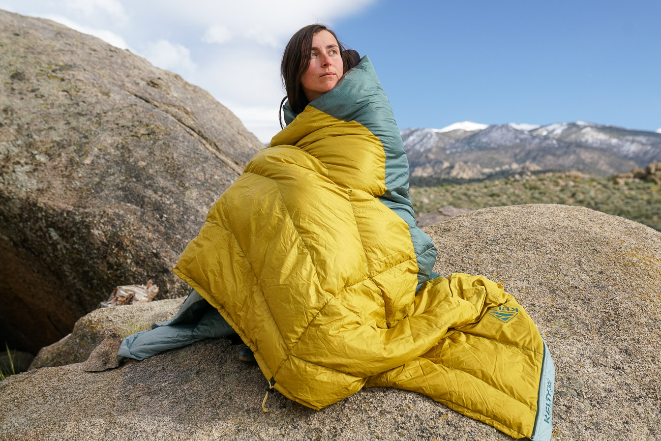 A person sits on a boulder wrapped in a sleeping bag.