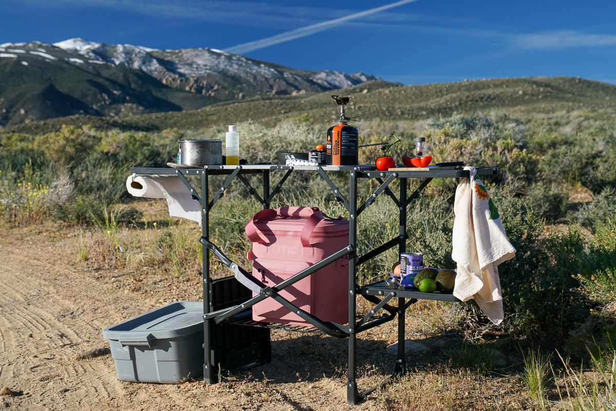A cook station loaded with ingredients and cooking items stands on the ground with mountains in the background.