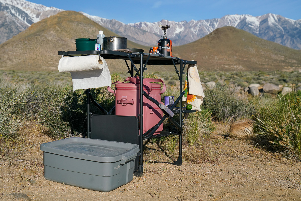 A cook station loaded with ingredients and cooking items stands on the ground with mountains in the background.