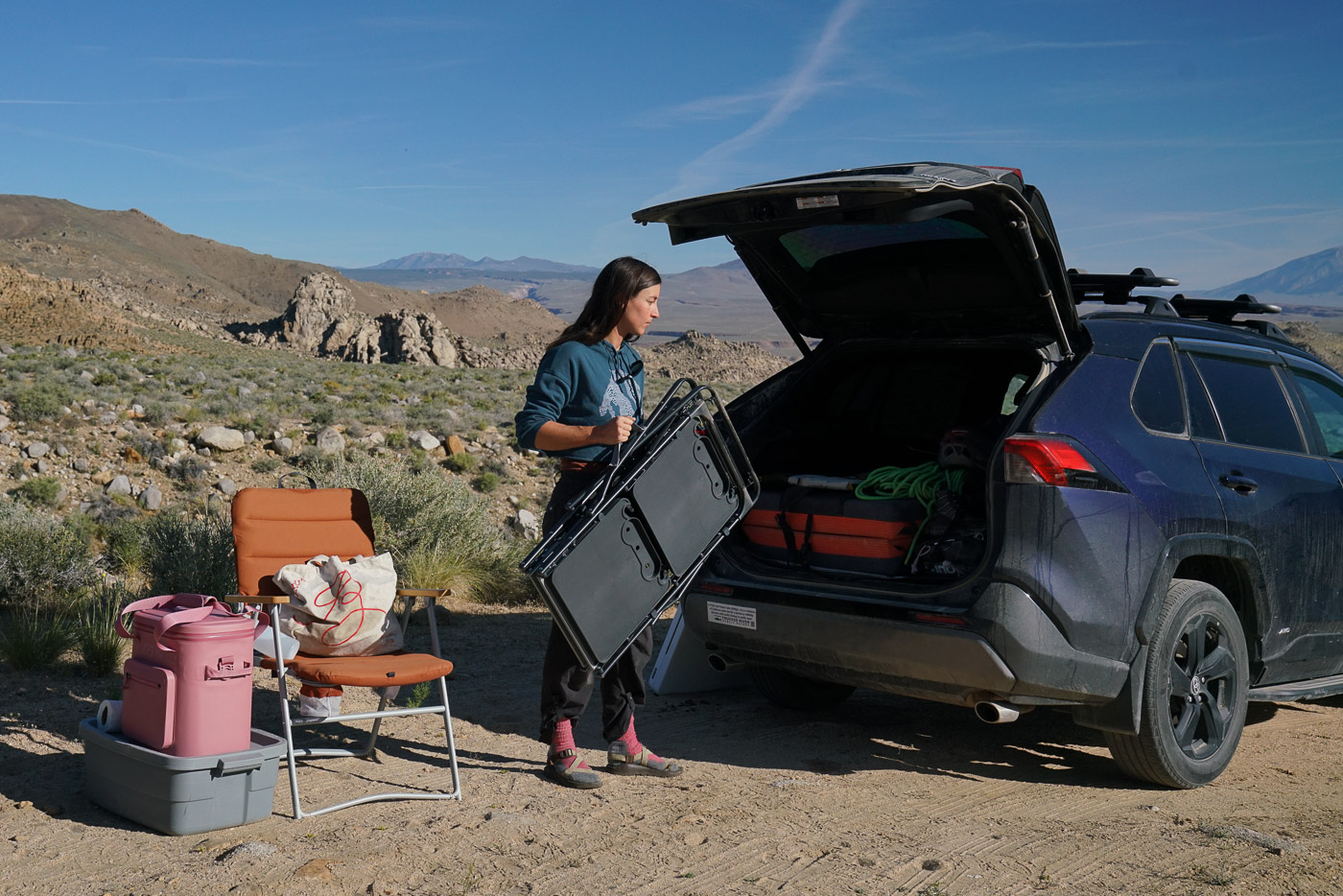 A person holds a folded cook station in front of the open trunk of a car. There are several items, including a cooler and a camp chair, next to them. There are large boulders and mountains in the background.