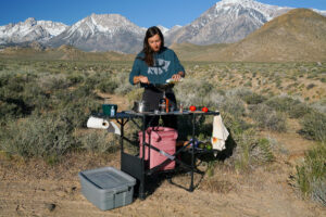 A person pours something into a frying pan sitting on top of a camp stove. The person stands in front of a cook station loaded with various kitchen items and ingredients. There are mountains in the background.