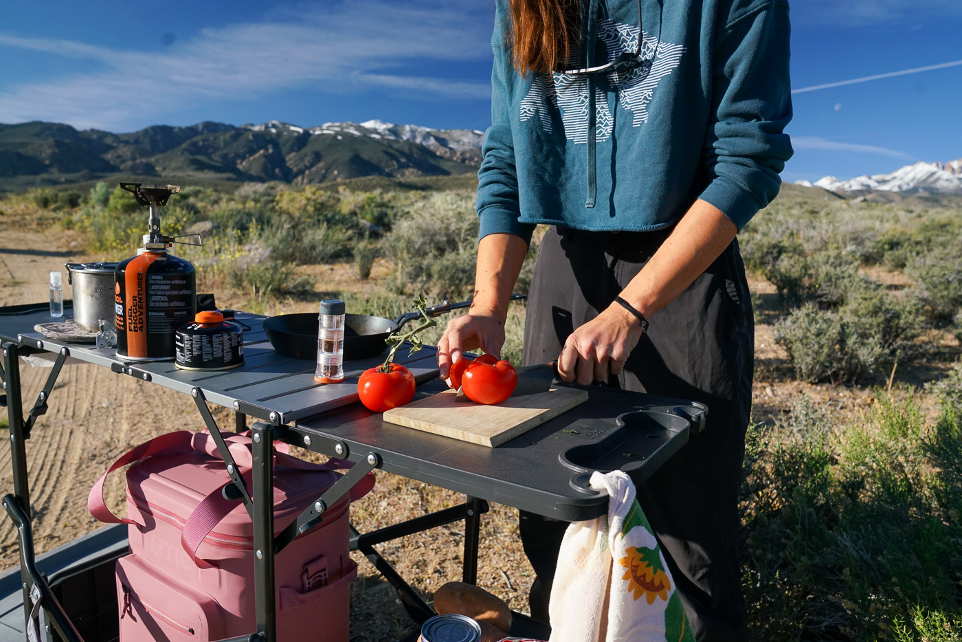 A close up of a person slicing a tomato in half on the shelf of a cook station. Beside them are various kitchen items.