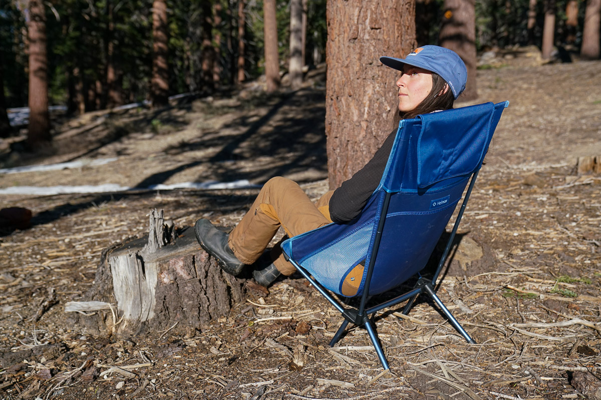 A person sits in a scoop-seat camping chair with their back to the camera in a pine forest with snow on the ground.
