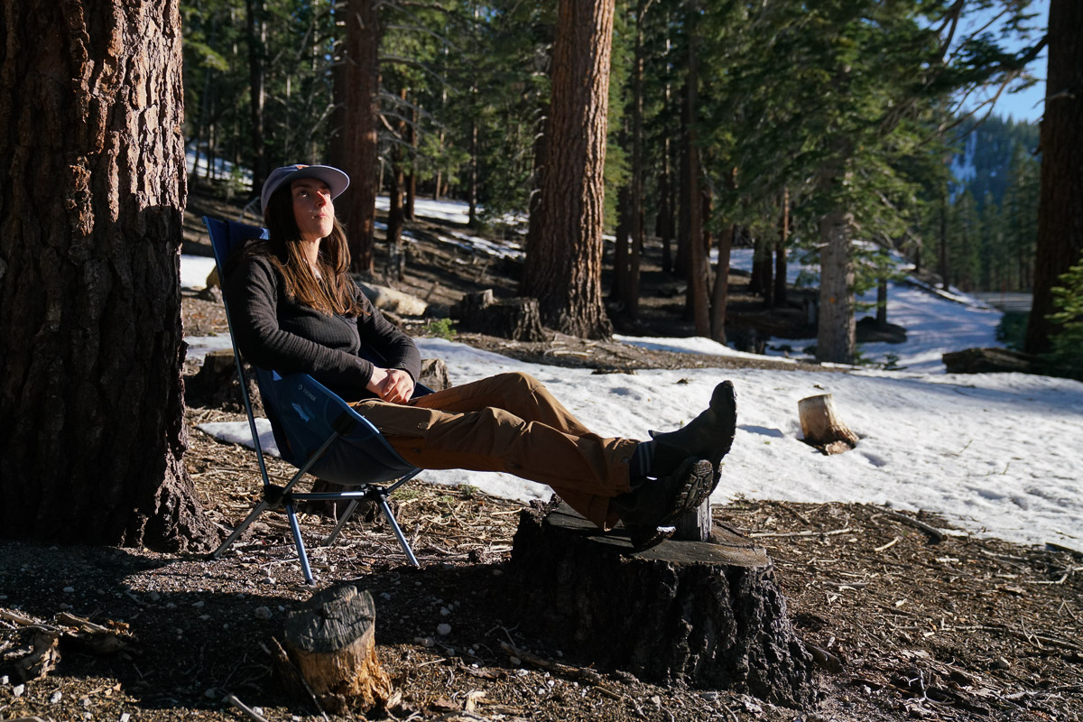 A person sits in a scoop-seat camping chair in a pine forest with snow on the ground.
