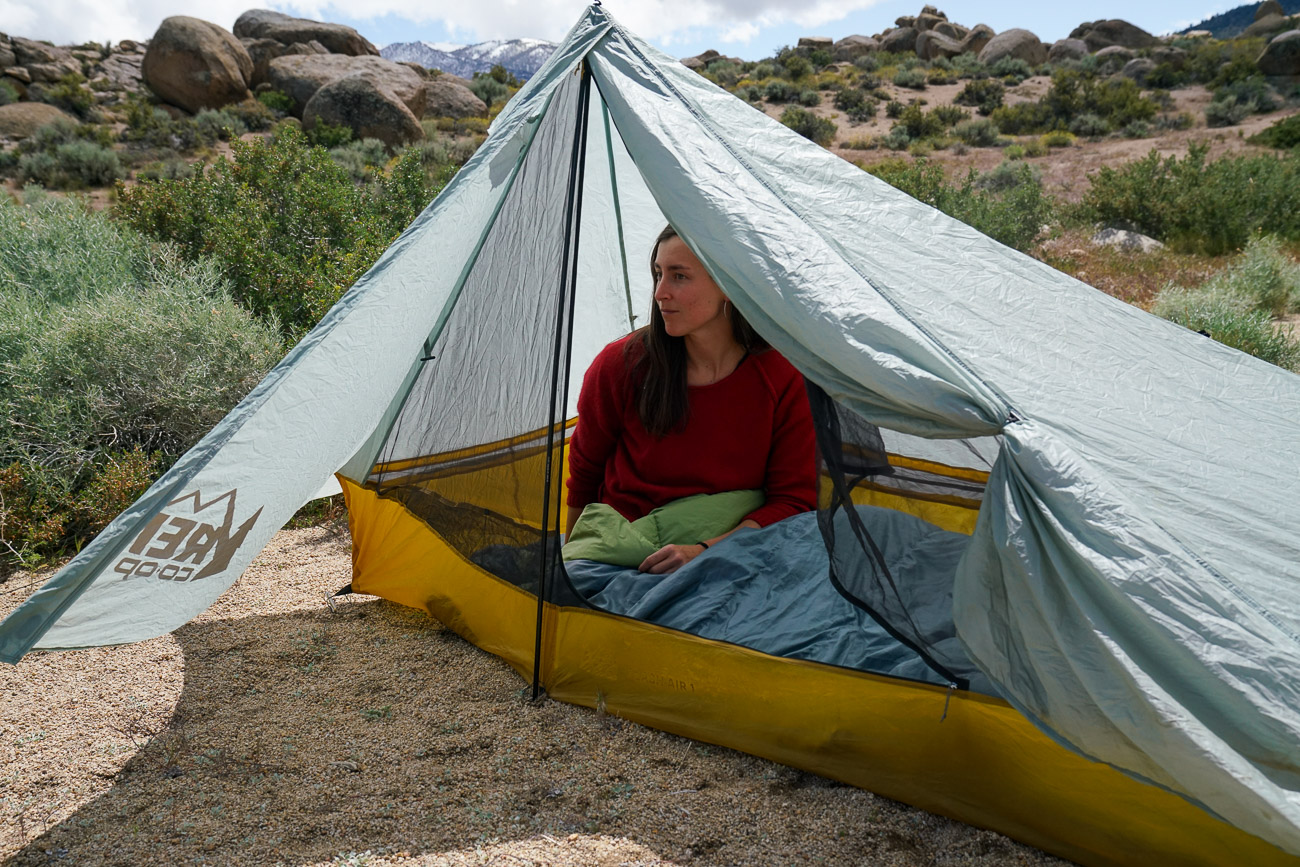 A person sits in a sleeping bag inside a tent with boulders in the background.