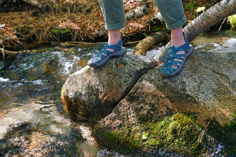 A close up of a person's feet wearing sandals standing on rocks in the middle of a creek.