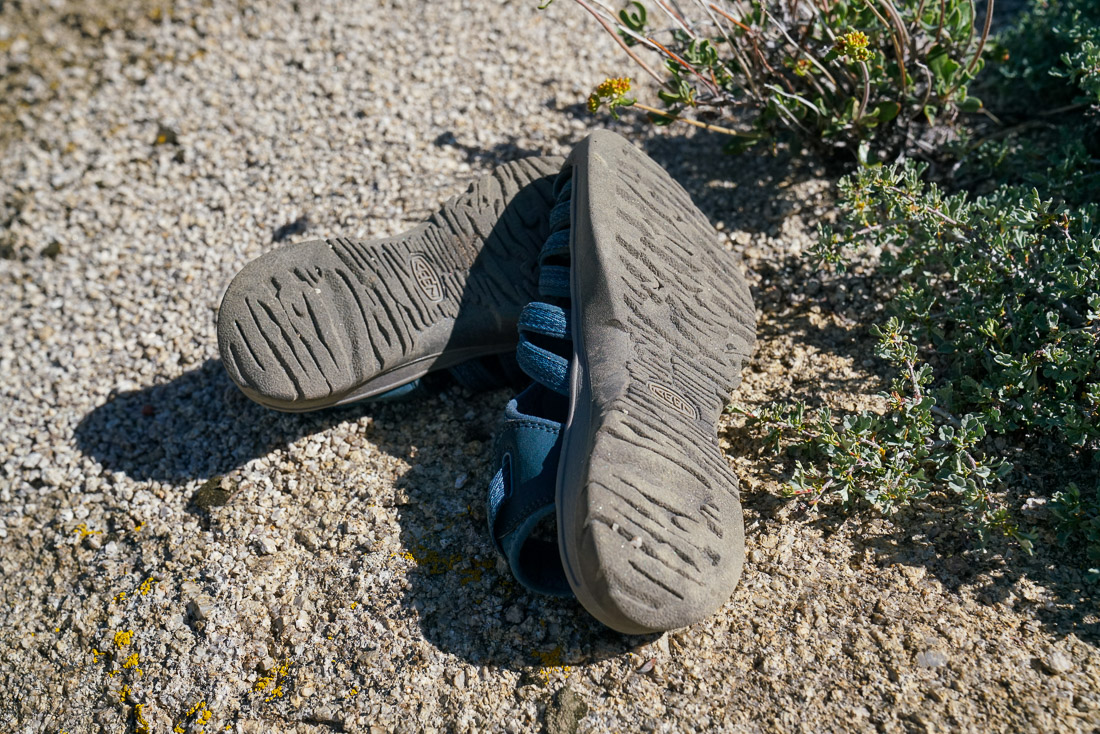 A close up of a pair of sandals flipped over to show their tread. The sandals rest on a rocky surface.