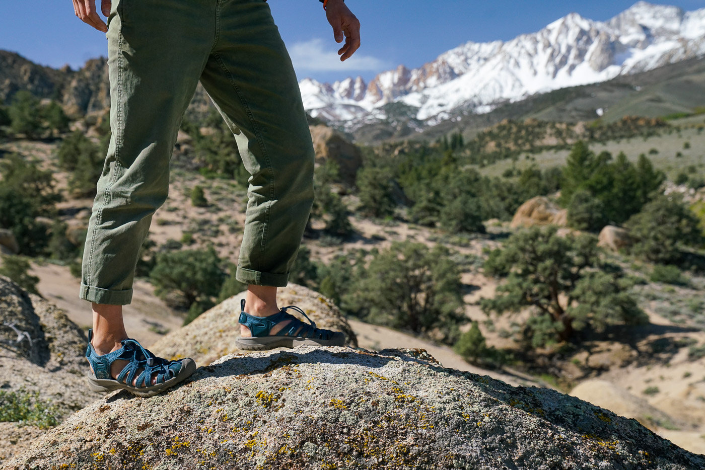 A person wearing sandals walks on a rocky surface with trees, boulders, and mountains in the background.