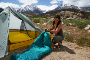 A person crouches on the ground and shoves a sleeping bag into a stuff sack. They kneel beside a tent with mountains in the background.