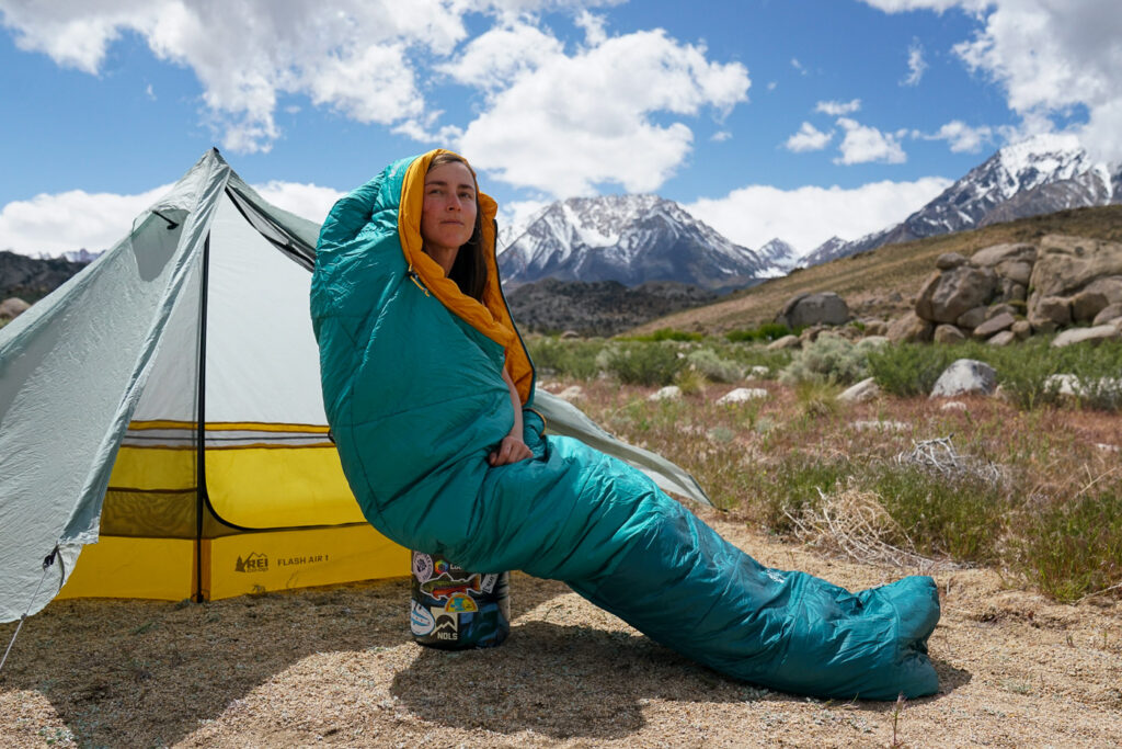 A person sits inside a sleeping bag on top of a bear canister. They sit in front of a tent with mountains in the background.