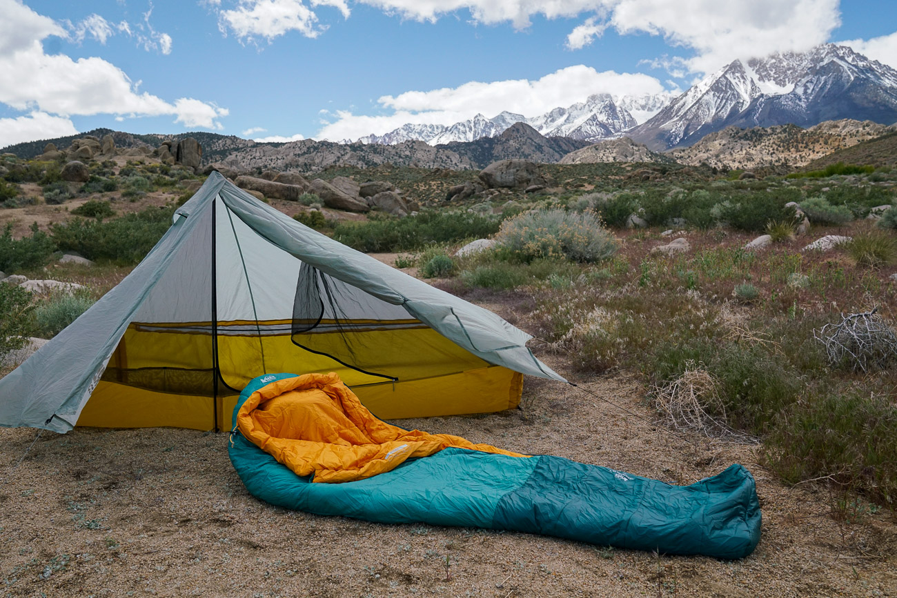 An empty sleeping bag lays on the ground outside a tent. There are mountains in the background.