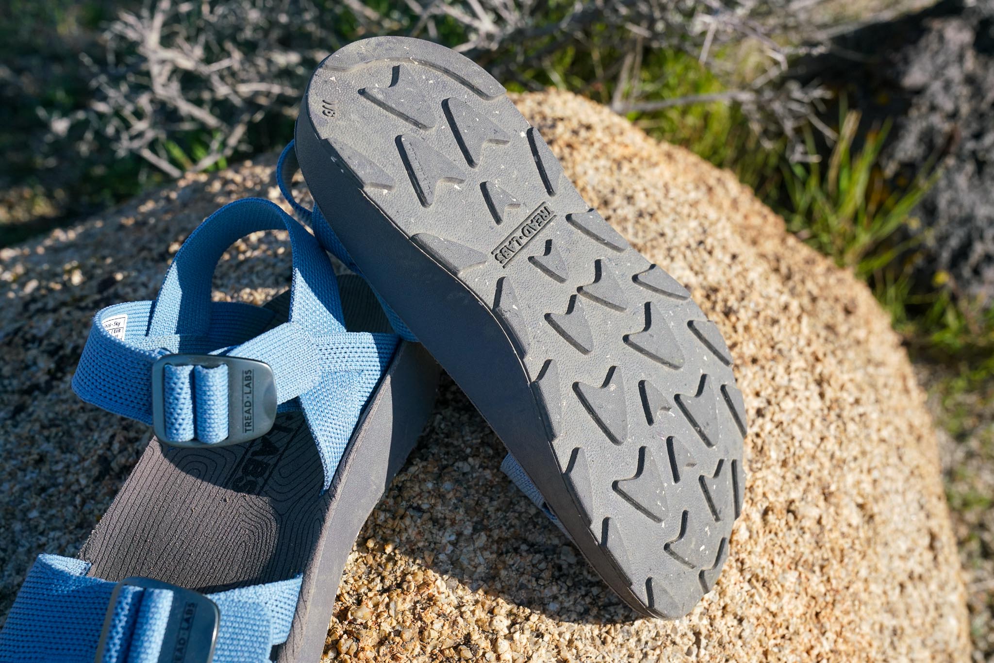 A pair of sandals rest on a rock with sagebrush in the background. One sandal is flipped over to show its bottom sole.
