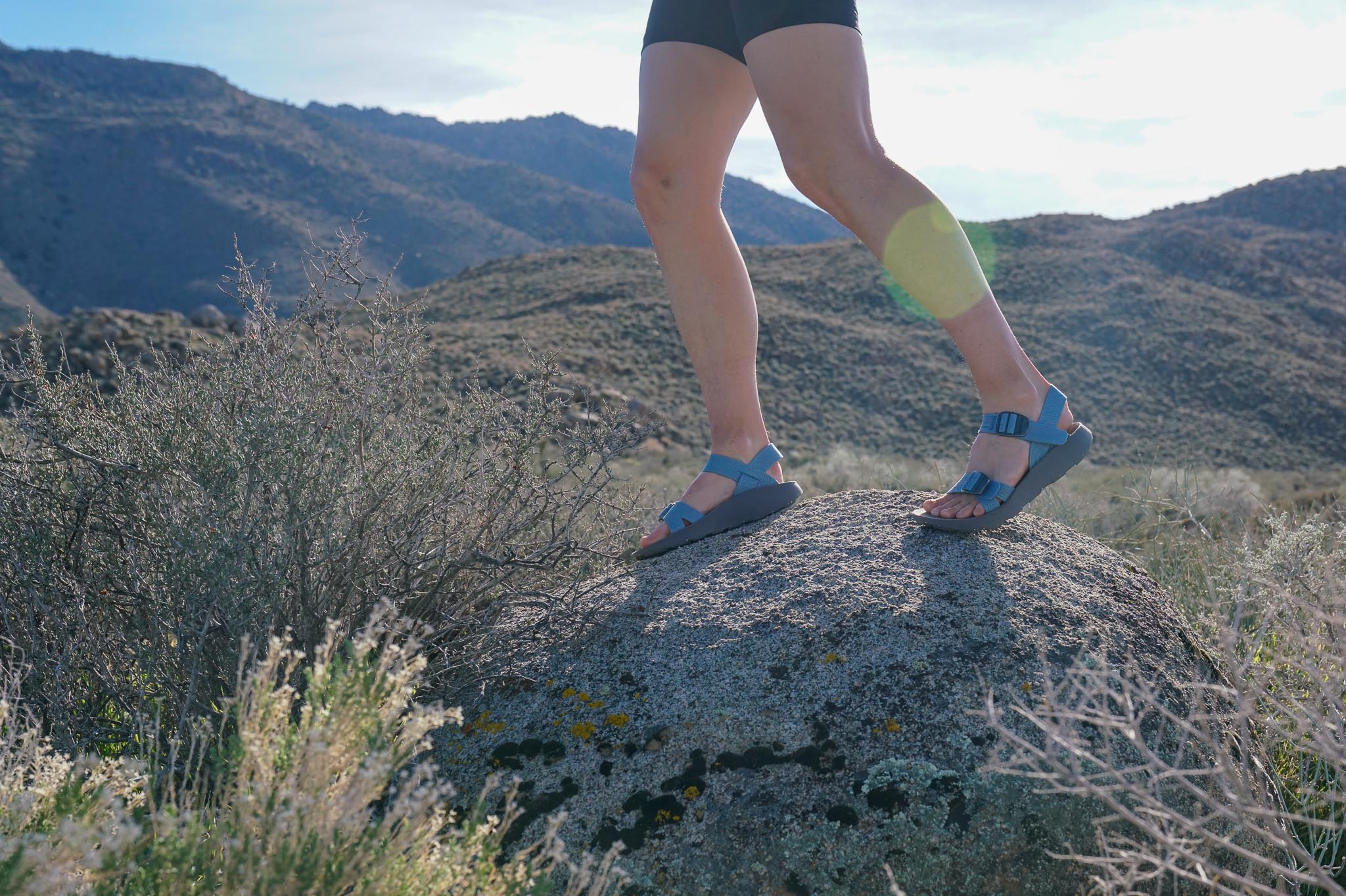 A close up of a person's feet wearing sandals standing on a rock with hills in the background.