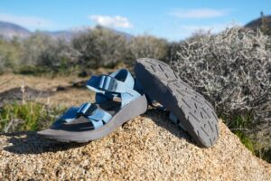 A pair of sandals rest on a rock with sagebrush in the background. One sandal is flipped over to show its bottom sole.