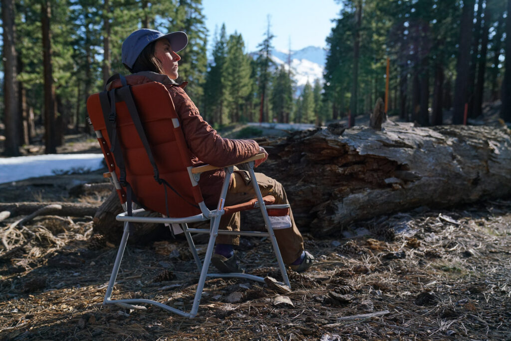 A person sits in a folding camping chair with their back to the camera with a pine forest in the background.