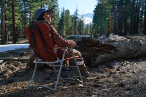 A person sits in a folding camping chair with their back to the camera with a pine forest in the background.