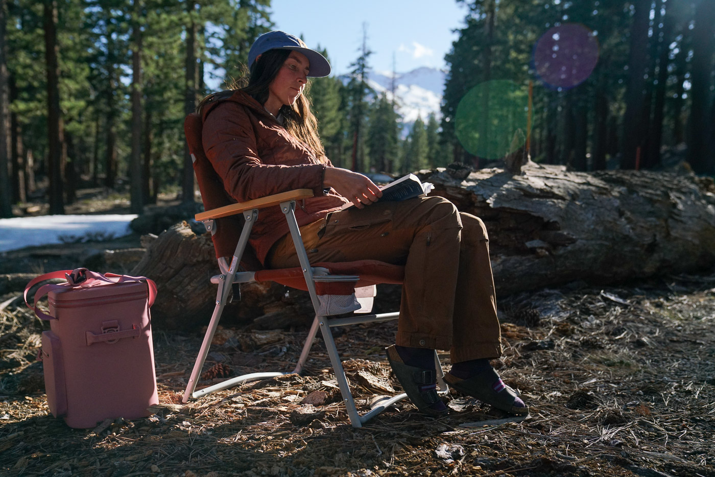 A person reads a book while sitting in a folding camping chair with a pine forest and mountains in the background.