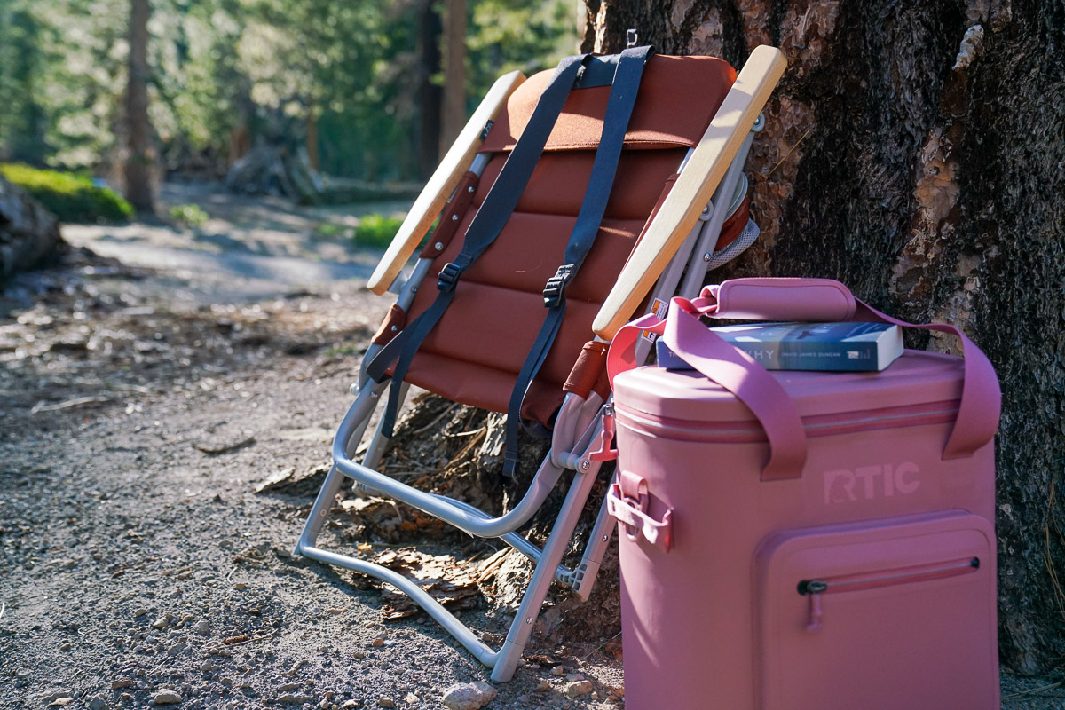 A folded camping chair rests against a tree with a small cooler beside it.