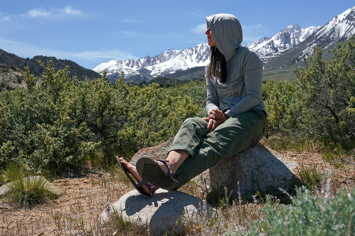 A person sits on a rock with their sandaled feet stretched in front of them. There are mountains in the background.