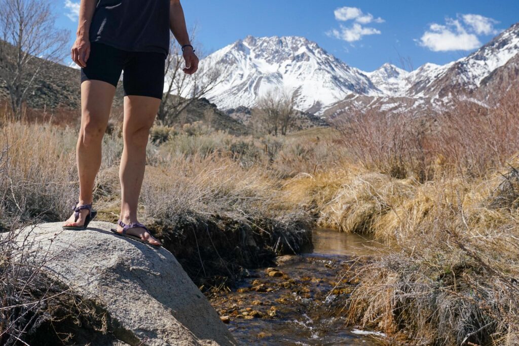 A person wearing sandals walks beside a creek with mountains in the background.