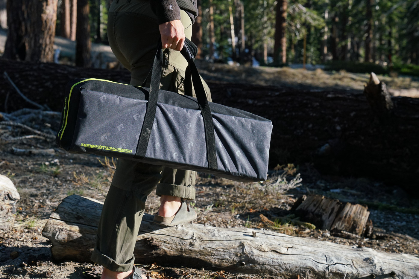 A close up of a person carrying a camp table in a case in a pine forest.