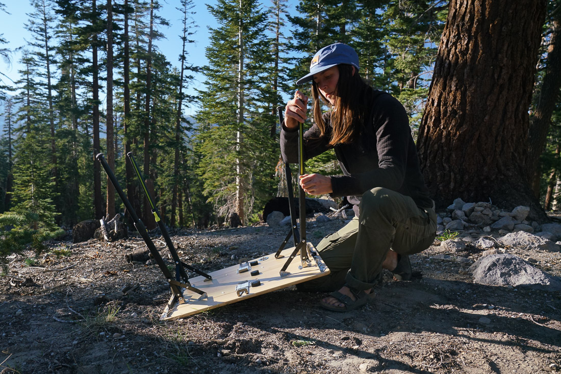A person extends the leg of a camping table with a pine forest in the background.