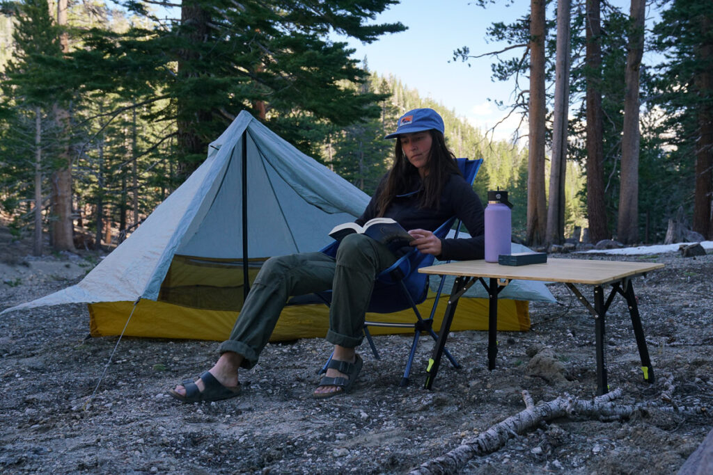 A person sit on a camp chair next to a camp table with a tent and pine forest in the background.