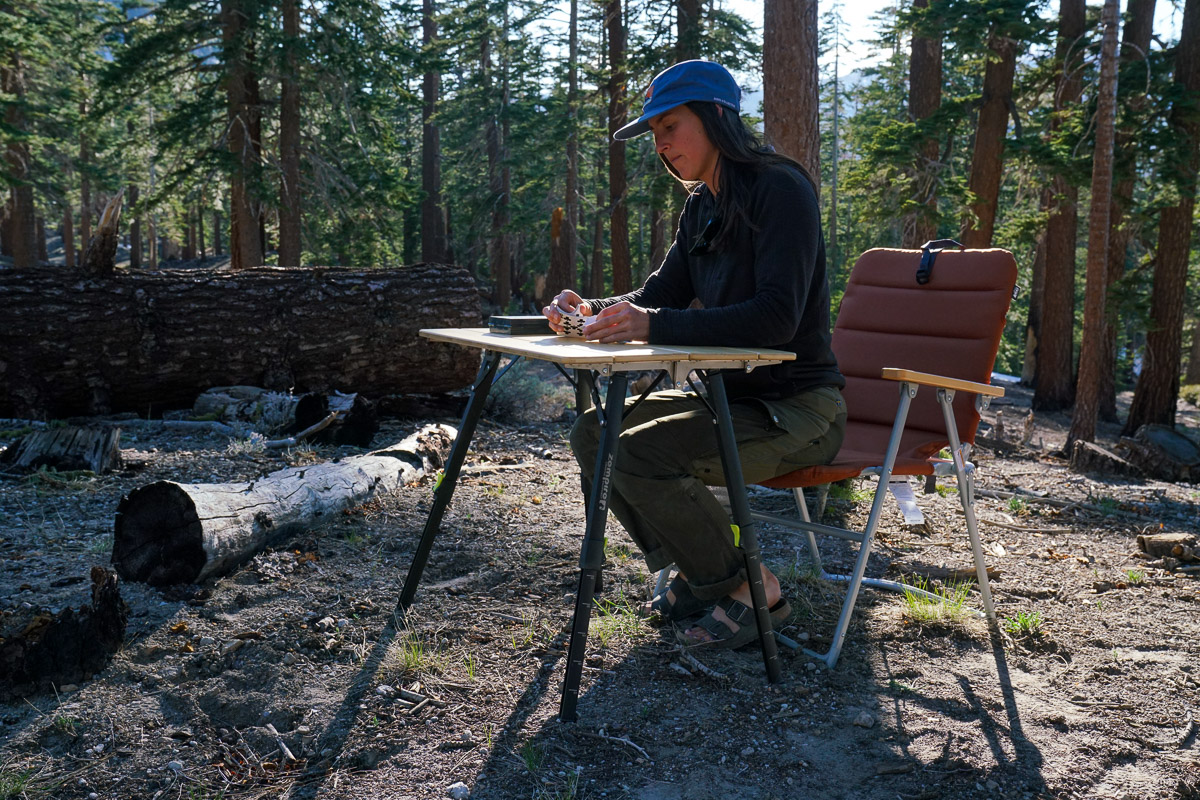 A person sits on a lawn chair at a camp table shuffling a deck of cards.