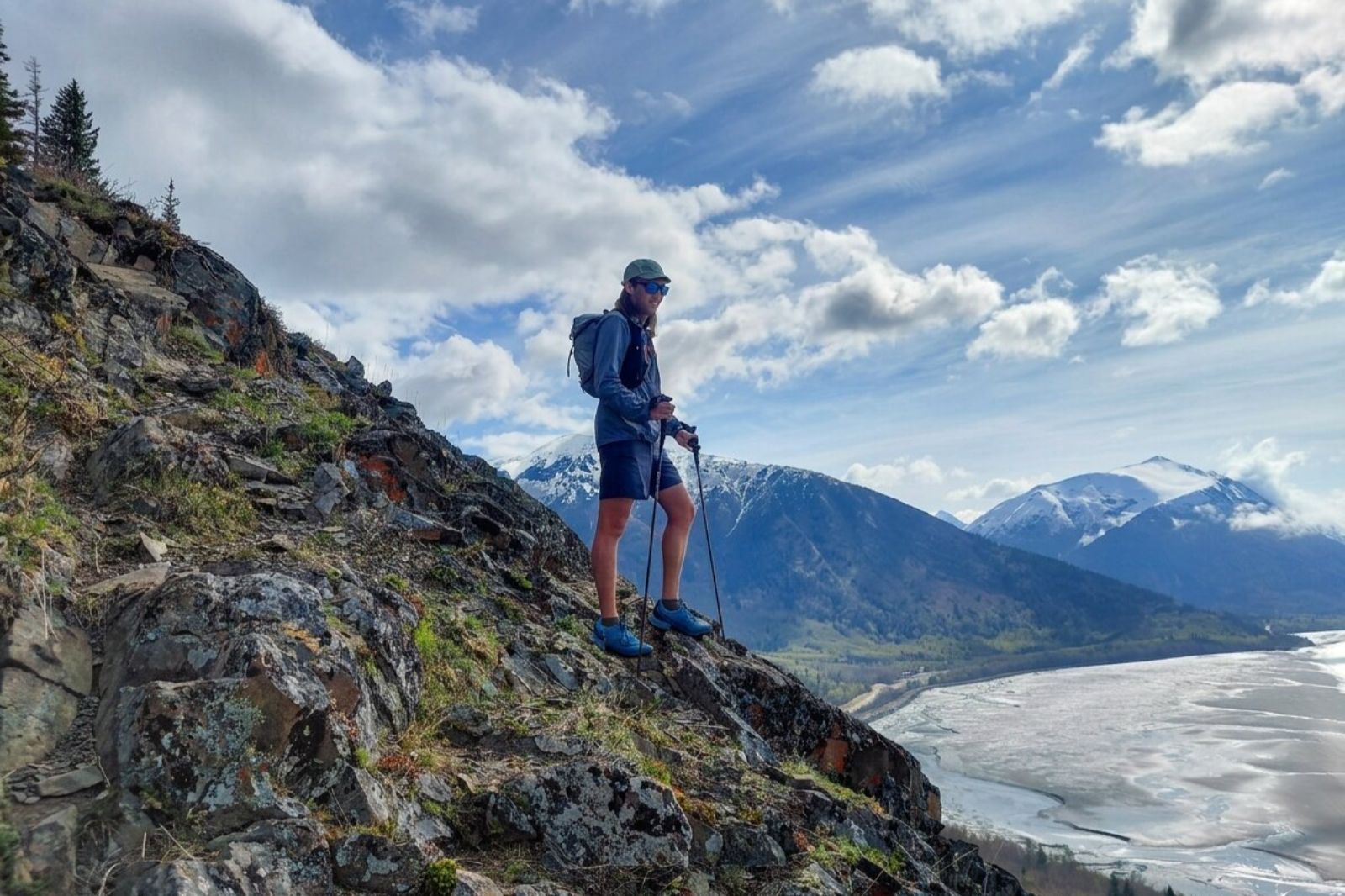 A hiker with trekking poles stands on a rocky hillside, overlooking mountains and the ocean.