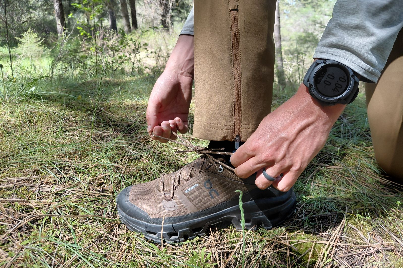 A person tying shoelaces on brown hiking shoes in a forested area.