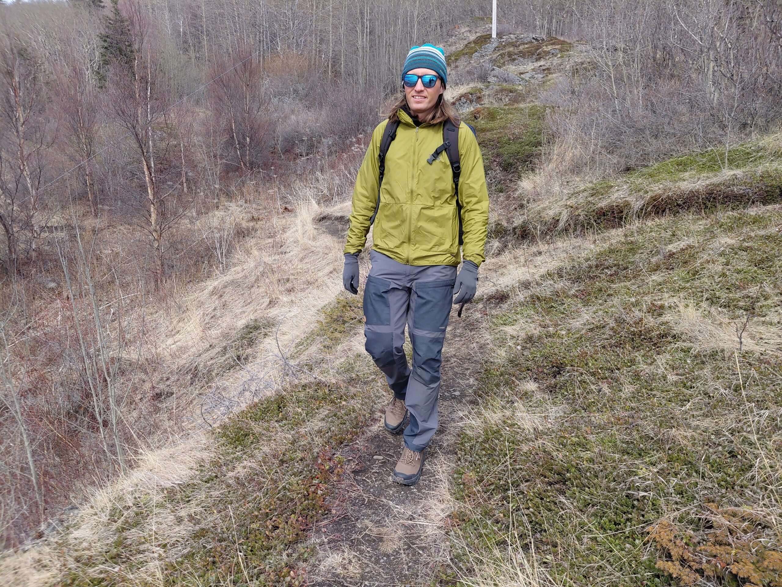 Person walking on a grassy trail in a sparse woodland, wearing outdoor gear.