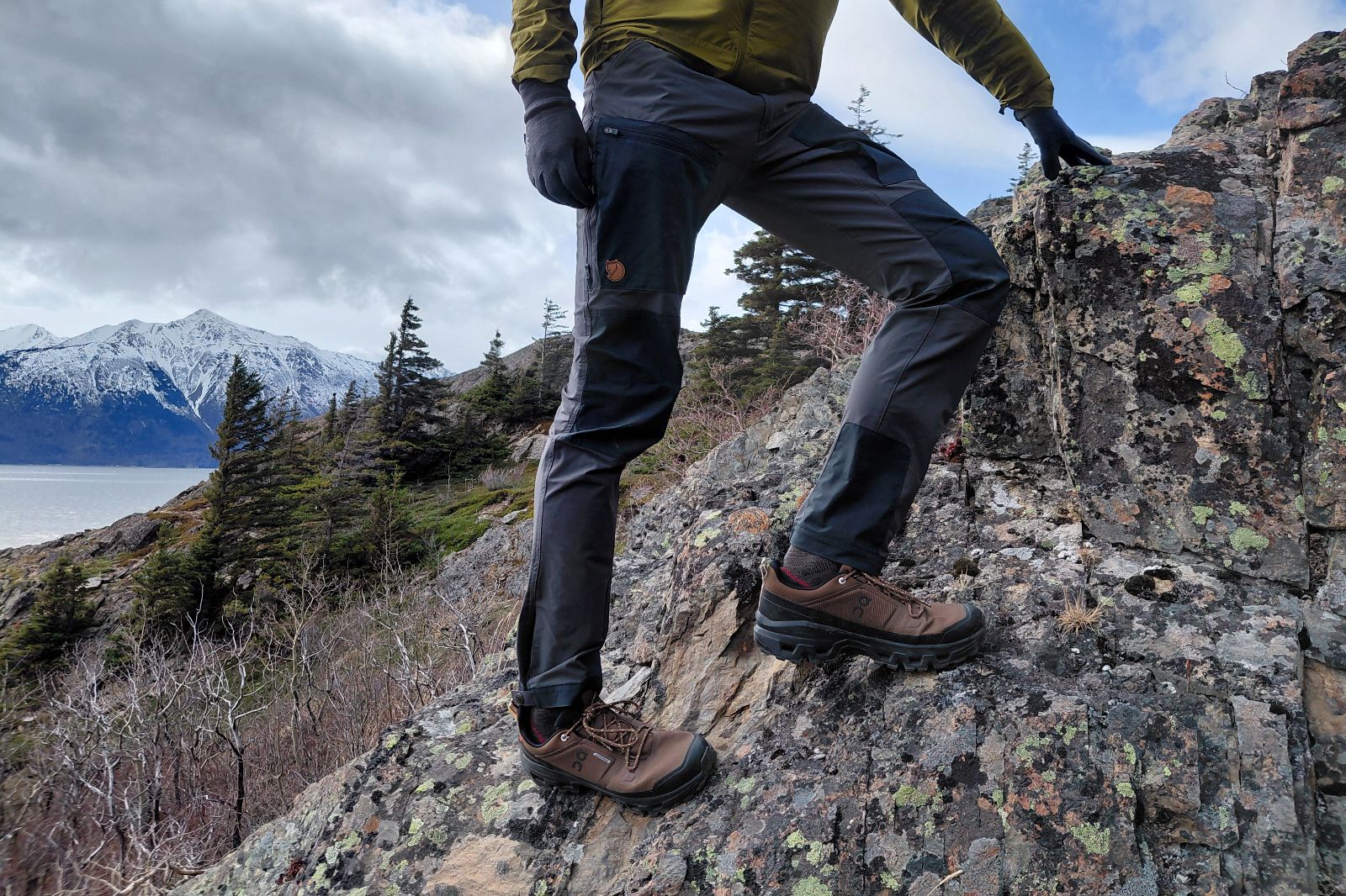 A hiker's legs and shoes on a rocky hillside with snow-capped mountains in the background.