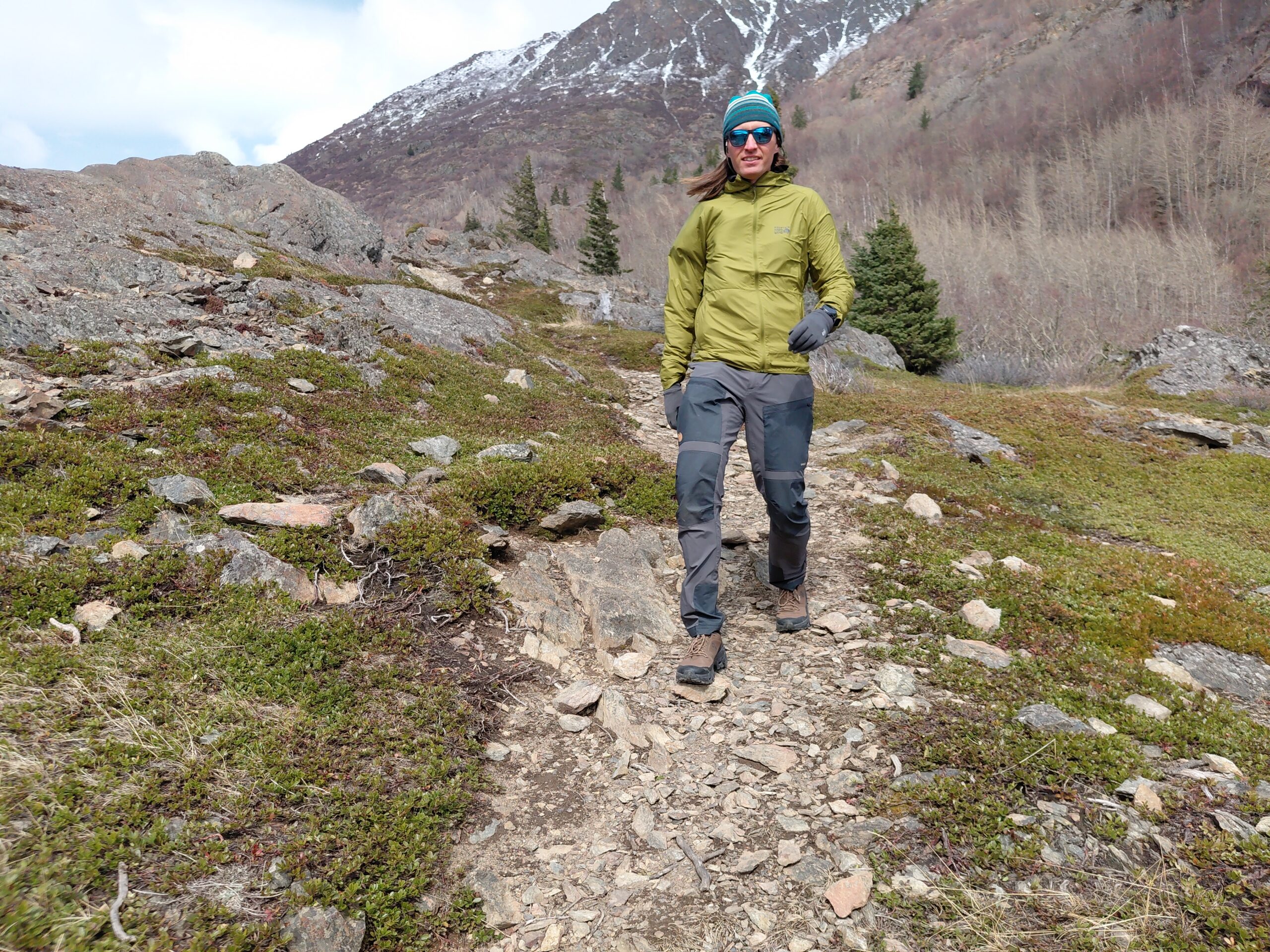Person hiking on a rocky mountain trail wearing a green jacket and beanie.