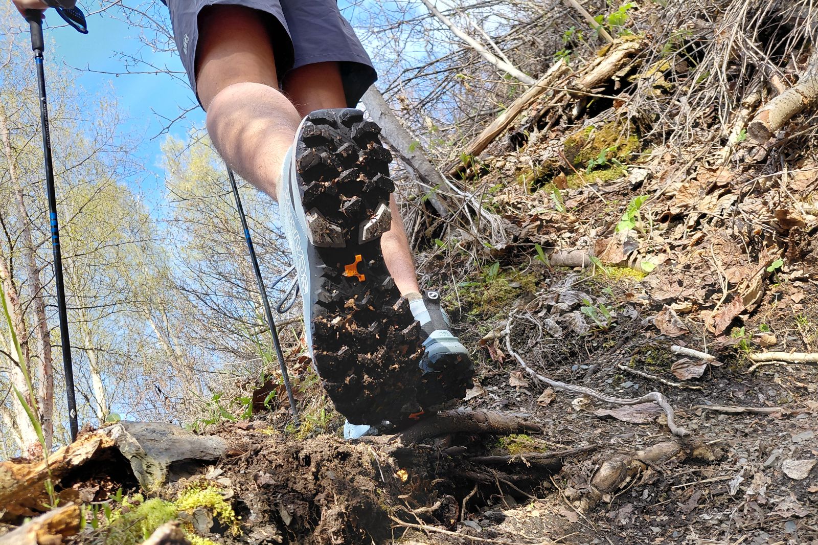 Close-up of a hiker's shoe tread as they walk on a rugged, leafy trail with trekking poles.