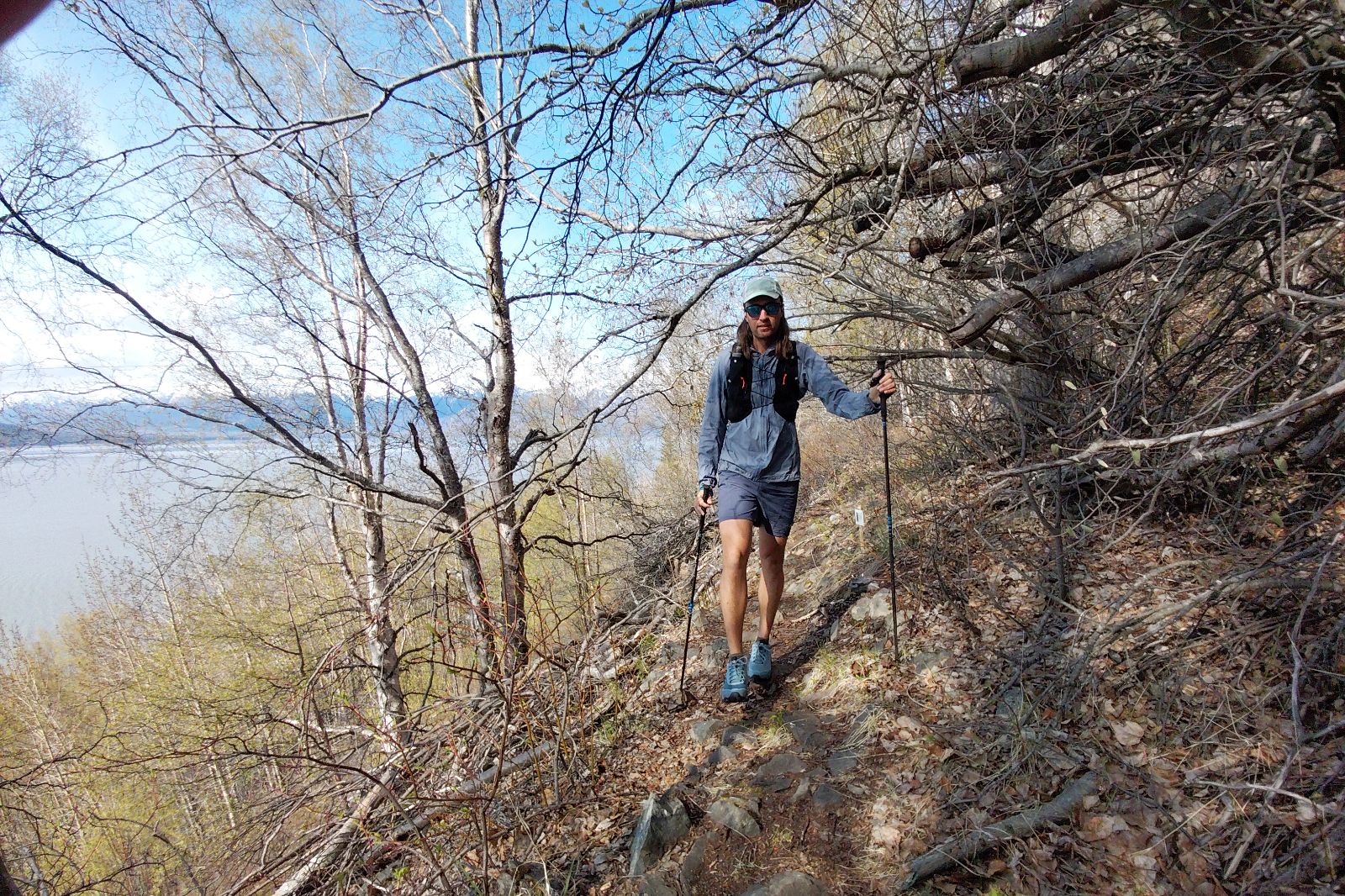 A hiker on a rocky forest trail with trekking poles, near a body of water and distant mountains.