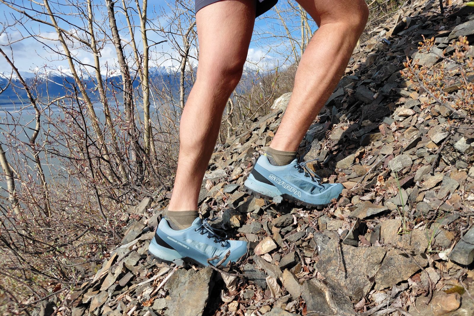 Close-up of a person in blue trail running shoes climbing a rocky slope, with mountains in the background.